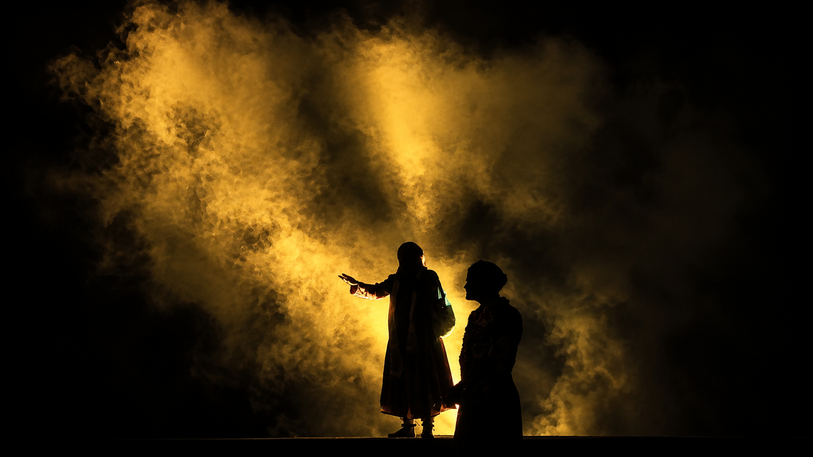 Two silhouetted performers surrounded by golden stage smoke